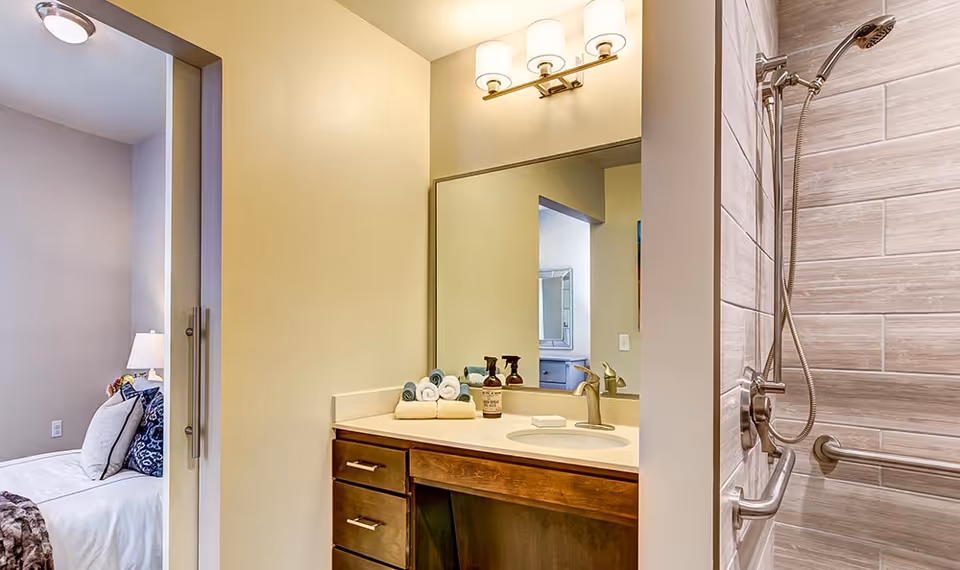 A bathroom with a wooden vanity featuring a white countertop and an oval sink. Above the sink is a large mirror with a three-light fixture. On the countertop are neatly rolled towels and soap dispensers. To the right is a tiled shower area with a handheld showerhead and a grab bar. Through an open doorway on the left, part of a bedroom with a bed, pillows, and a lamp is visible.