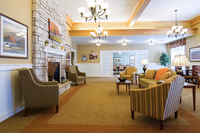 A spacious and well-lit living room area in an assisted living facility featuring comfortable armchairs and sofas arranged around a coffee table. The room has a stone fireplace with a painting above it, multiple chandeliers hanging from the ceiling, and framed artwork on the walls. There is a dining area visible in the background through glass-paneled double doors.