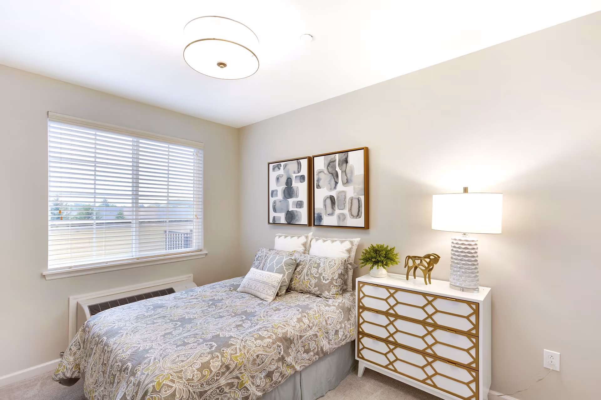 A neatly made bed with patterned bedding in a bright bedroom featuring a window with blinds, two abstract paintings on the wall above the bed, and a white dresser with gold geometric accents. A table lamp and a small plant are placed on the dresser.