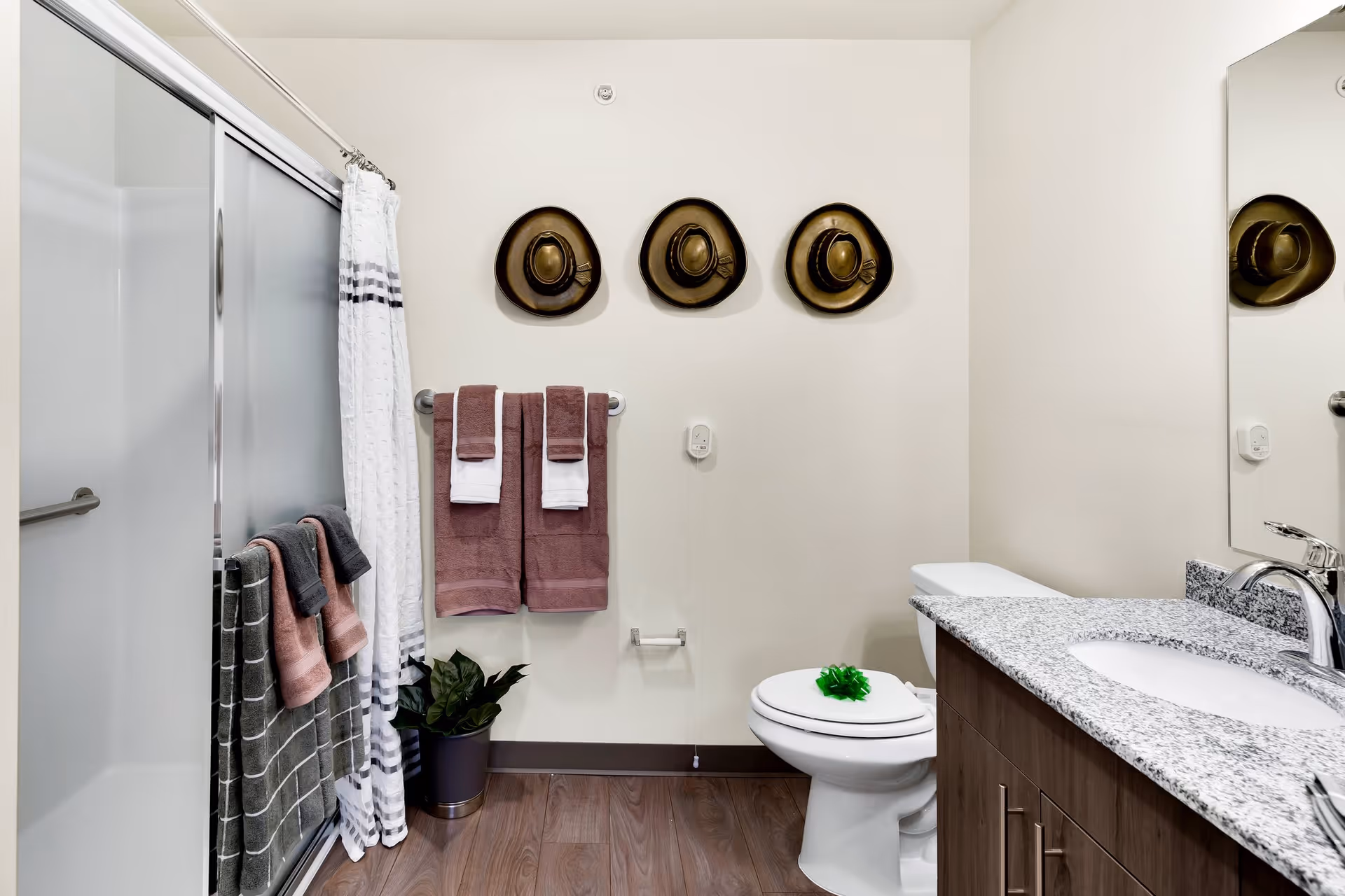 A clean and modern bathroom featuring a shower with sliding glass doors and a white and gray striped shower curtain. There are several towels hanging on racks, including brown and white towels. Three decorative hats are mounted on the wall above the towel rack. A white toilet with a green decorative item on the lid is next to a wooden vanity with a granite countertop and a sink. A small potted plant is placed on the floor near the shower.