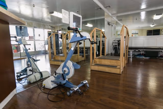 A rehabilitation or physical therapy room with a stationary exercise bike, wooden stairs with handrails for physical therapy exercises, a treatment table, and large mirrors on the wall reflecting the equipment and room.