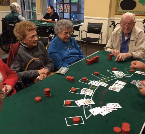 A group of elderly people sitting around a green felt card table playing a card game with red poker chips. There are three elderly individuals prominently visible, two women and one man, engaged in the game. In the background, other people are seated and interacting in a well-lit room with windows and colorful decorations on the walls.