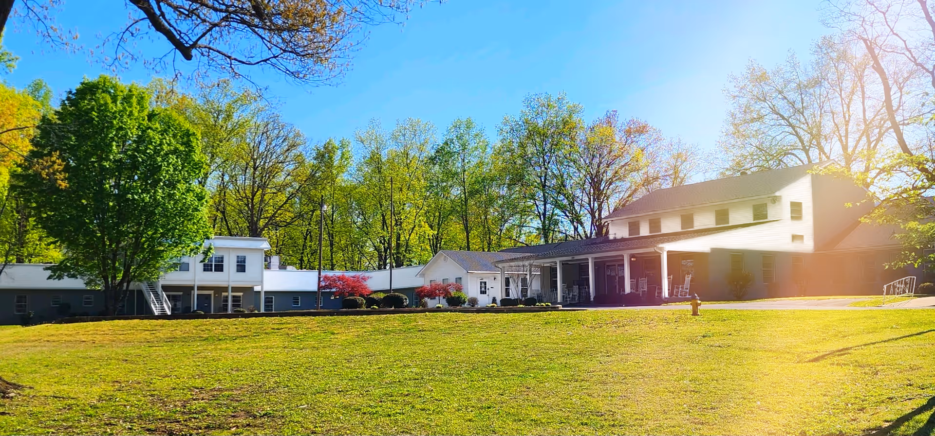 White two-story senior living building with a covered porch and an expansive grassy lawn under a sunny blue sky.
