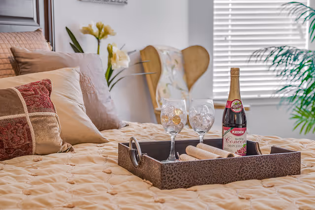 A cozy bedroom with a neatly made bed featuring a beige quilted bedspread and multiple decorative pillows in shades of beige and red. On the bed is a dark tray holding two empty wine glasses and a bottle of sparkling Concord grape juice. In the background, there is a wooden chair with a floral cushion, a window with white blinds, and a green plant.