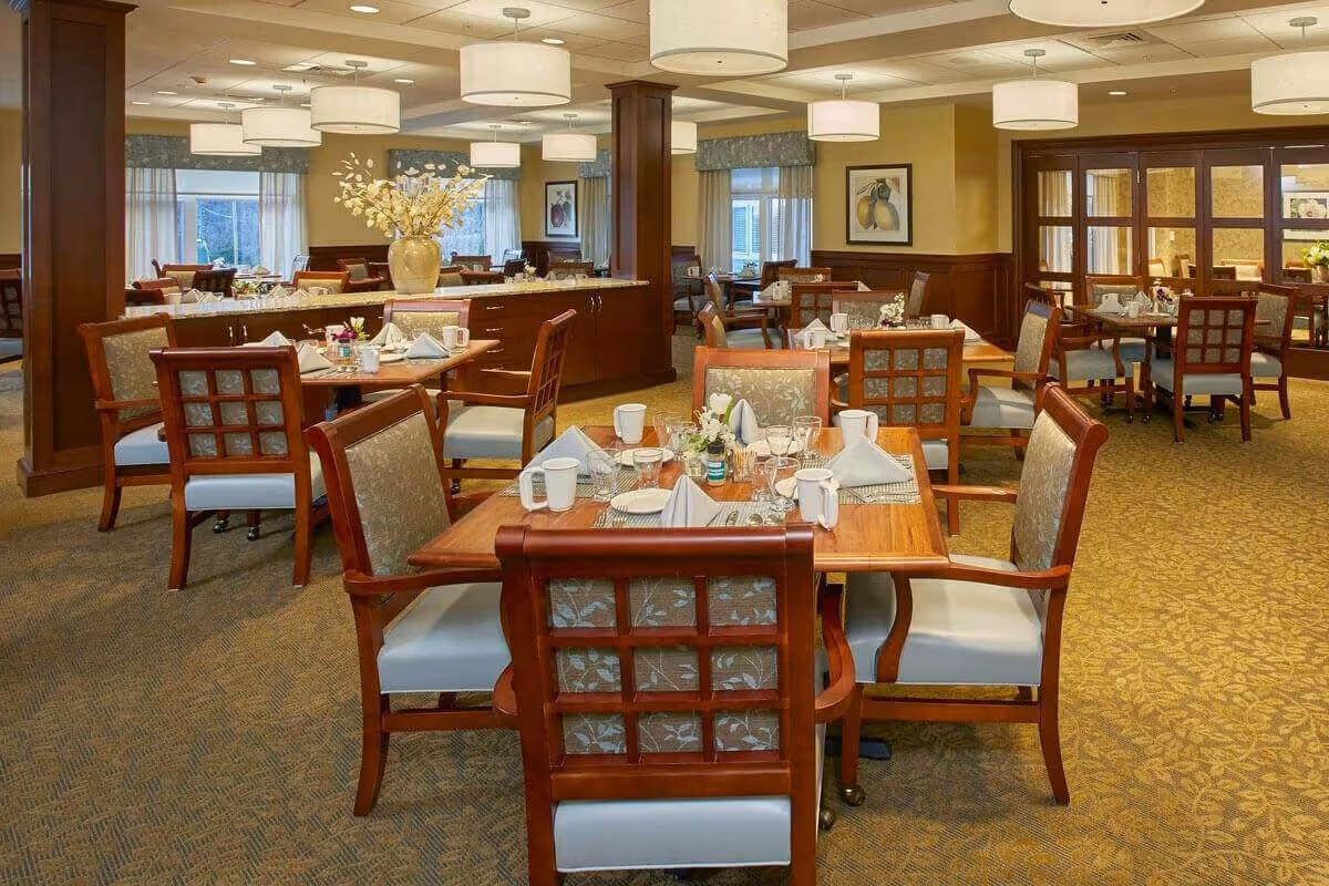 A well-lit dining room in a senior living facility with several wooden tables and cushioned chairs arranged neatly. Each table is set with white napkins, cups, glasses, and small flower arrangements. The room features warm beige walls, carpeted floors with a patterned design, large windows with curtains, and multiple ceiling light fixtures. There are framed fruit-themed paintings on the walls and a large vase with flowers on a central counter.
