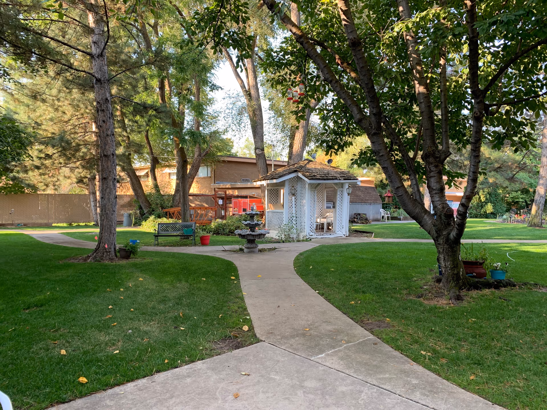 A peaceful outdoor garden area at Williamsburg Retirement Community featuring a paved walkway that splits into two paths, green grass, several trees, a small white gazebo, a multi-tiered fountain, and benches surrounded by potted plants.