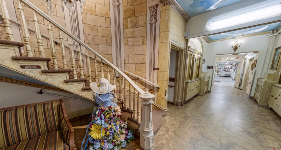Interior hallway of Hughes Health & Rehabilitation featuring a curved staircase with ornate railing and a mannequin dressed in a floral dress and wide-brimmed hat seated on a striped bench. The hallway has decorative wall panels, a painted ceiling, and light fixtures along the corridor.