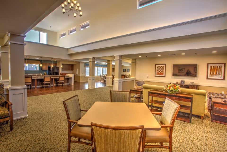 A spacious and well-lit common area in a senior living facility with a high ceiling and large windows near the ceiling. The room features a dining table with four chairs in the foreground, a seating area with sofas and armchairs, a flat-screen TV mounted on the wall, and a kitchen area with bar stools and pendant lighting in the background. The decor includes framed artwork on the walls and a floral centerpiece on a console table behind the sofa.