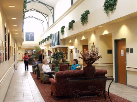Bright interior common lounge of a senior living community with sofas, plants, a vaulted skylight and residents chatting.