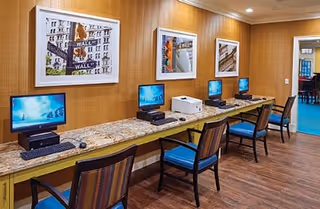 A computer workstation area with four desktop computers and chairs along a long marble countertop against a wood-paneled wall. Three framed pictures hang on the wall above the computers. The floor is wooden, and there is a doorway leading to another room with blue carpet.