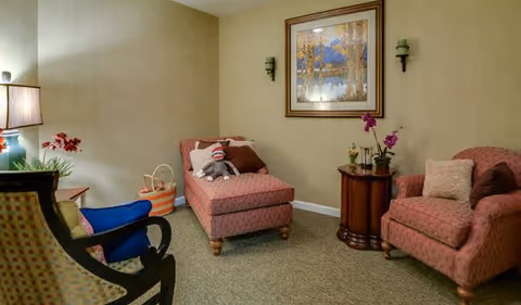 A cozy living room area with a patterned chaise lounge and matching armchair, a small round wooden table with potted orchids, a framed painting on the wall, and a basket with a stuffed sock monkey toy. The room has beige walls and carpeted floor, with a table lamp and additional seating visible.