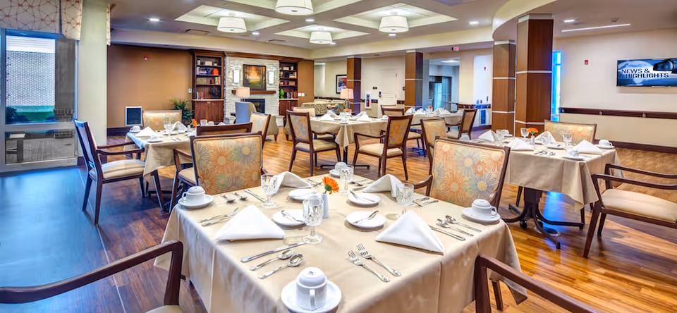 A well-lit dining room in a senior living facility with multiple tables covered in beige tablecloths, each set with white napkins, plates, cups, and silverware. The chairs have floral patterned upholstery. The room features wooden flooring, a fireplace with a mirror above it, built-in bookshelves, and a wall-mounted TV displaying 'News & Highlights'.