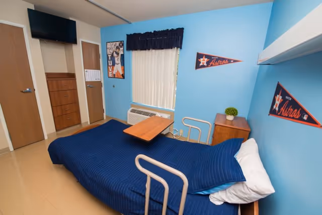 A small bedroom in a care facility with a single bed covered in a blue blanket, a wooden bedside table with a small plant, and a window with vertical blinds and a dark valance. The walls are painted blue and decorated with Houston Astros pennants and a sports poster. There is a mounted TV above a set of drawers and two closed wooden doors.