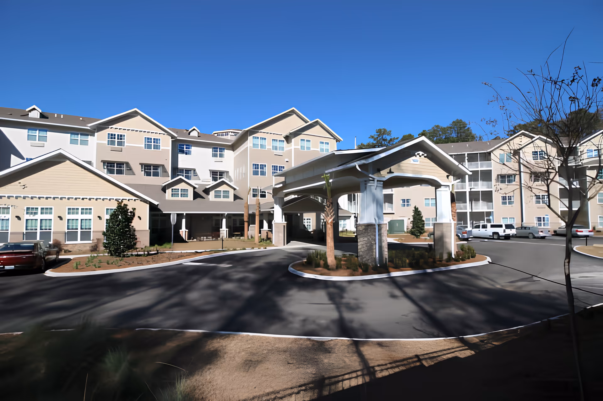 Exterior view of Mulligan Park Gracious Retirement Living facility showing a multi-story building with beige and white walls, numerous windows, a covered driveway entrance, parked cars, and clear blue sky.