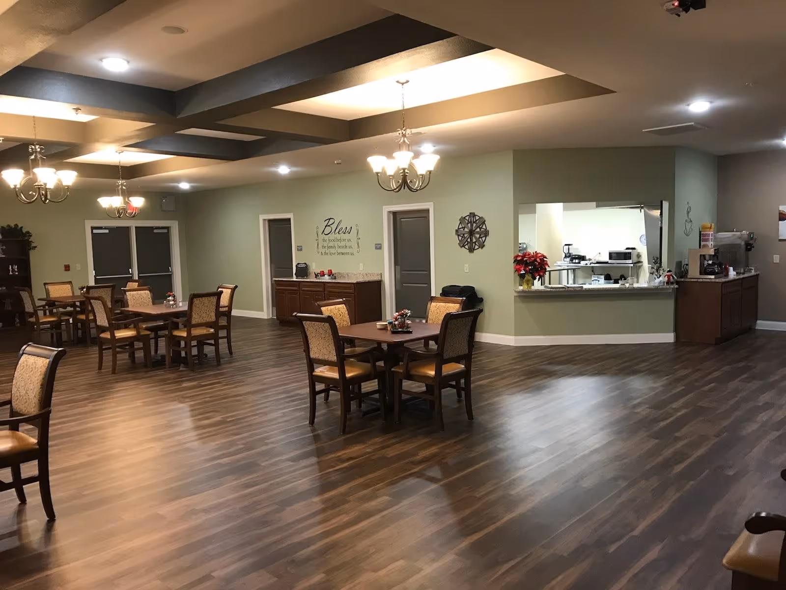 A spacious dining area in a retirement village with several wooden tables and chairs arranged on a wood-patterned floor. The ceiling features recessed lighting and chandeliers. There is a serving counter with a kitchen visible behind it, and a wall with a decorative clock and a sign that reads 'Bless the food before us, the family beside us, & the love between us.'