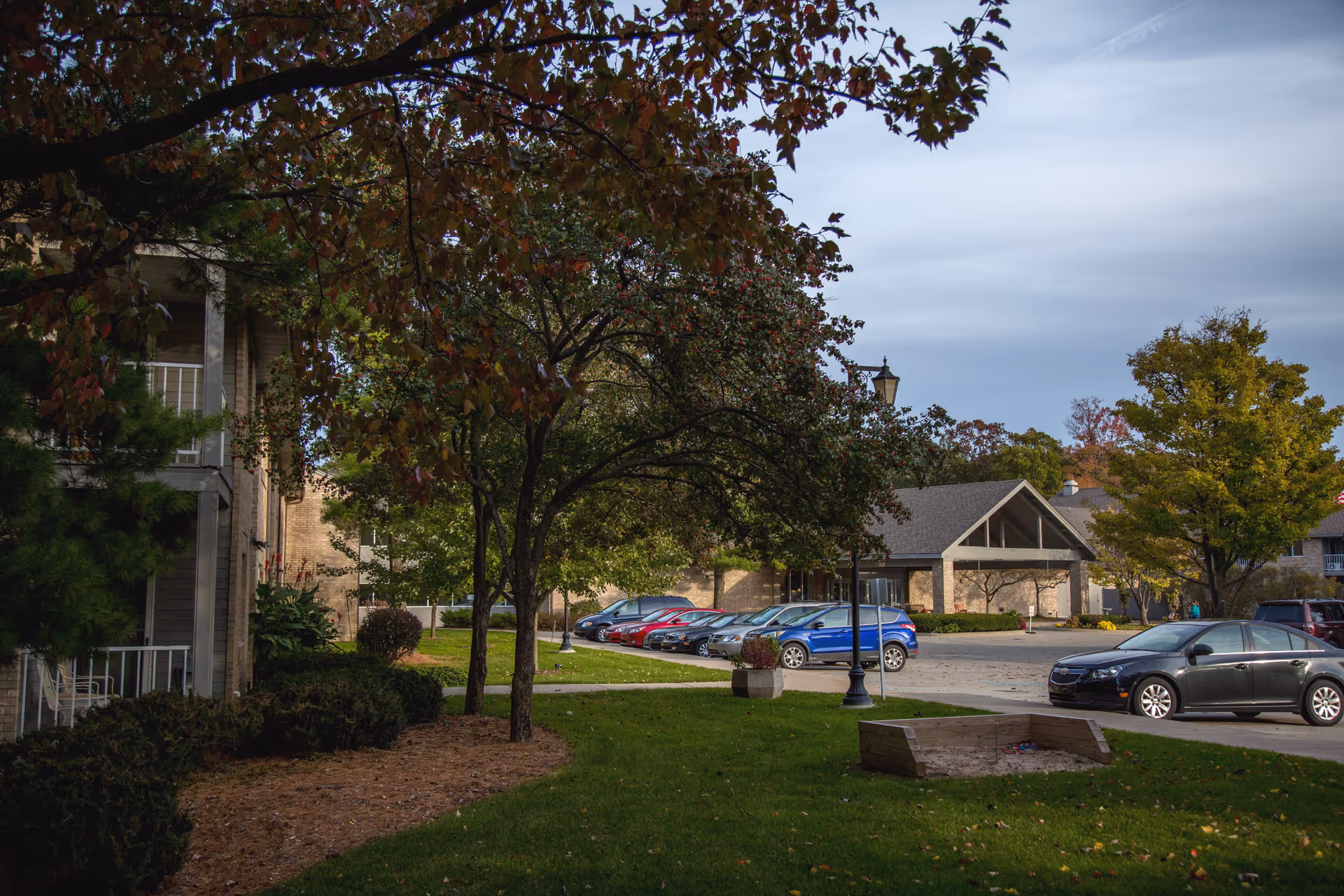 Outdoor view of Sentinel Pointe Retirement Community showing a parking lot with several cars, trees with autumn foliage, a grassy area with a wooden sandbox, and parts of the building in the background under a cloudy sky.