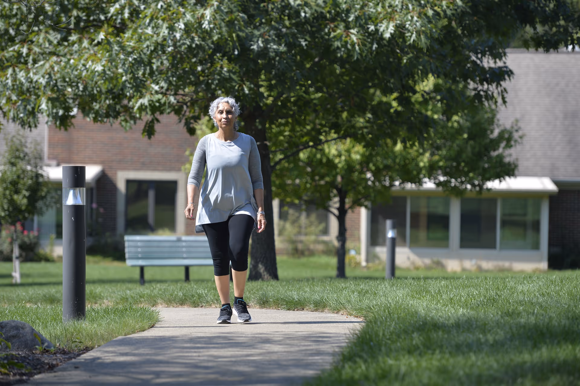 An older woman with gray hair wearing a light gray long-sleeve top and black leggings walking on a paved path in a green outdoor area with trees, grass, and a bench, with a building visible in the background.