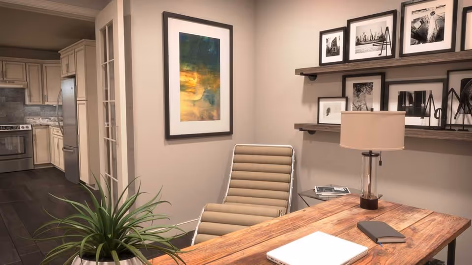 A cozy interior space featuring a wooden desk with a closed laptop, a notebook, and a lamp. Behind the desk is a modern beige chair. The wall has two wooden shelves displaying framed black and white photographs, and a colorful abstract painting is hung on the adjacent wall. In the background, a kitchen area with light-colored cabinets, a stainless steel refrigerator, and a stove is visible. A green potted plant is placed near the desk.