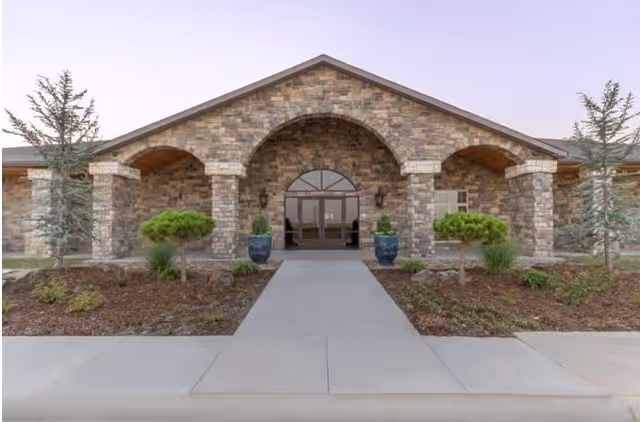 Front exterior view of a stone building with a peaked roof and three archways at the entrance. There are two large blue planters with greenery on either side of the glass double doors. Small trees and shrubs are planted in landscaped beds along the walkway leading to the entrance.