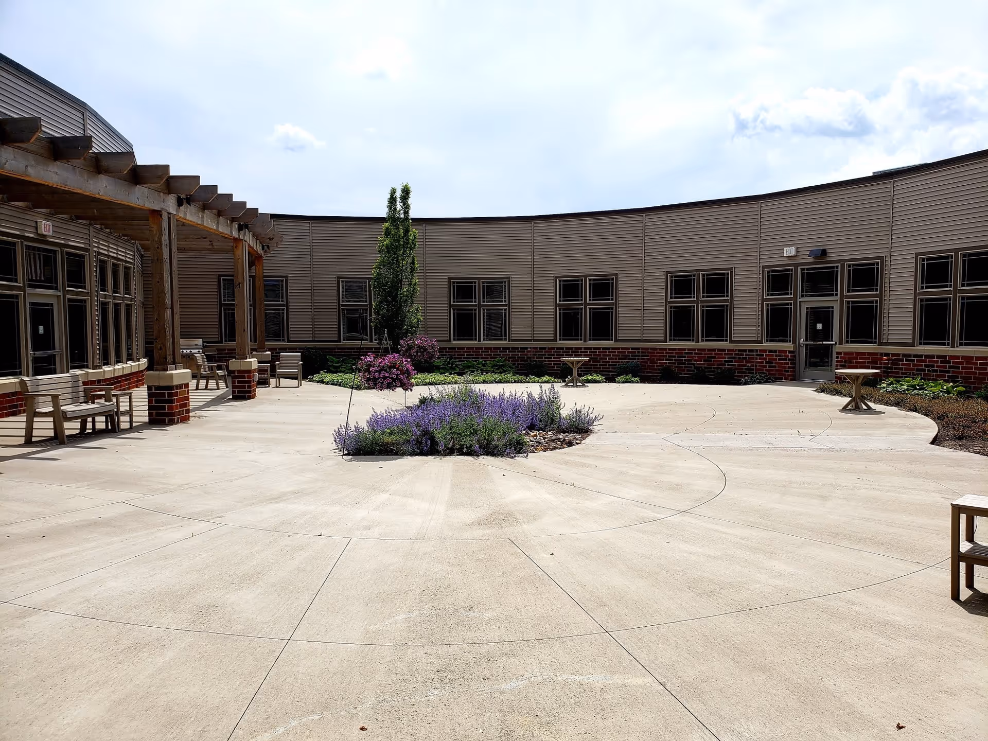 Outdoor courtyard area at The Bridges at Ankeny featuring a circular concrete patio with a central flower bed of purple flowers and greenery. Surrounding the patio are beige siding walls with multiple windows and doors, some wooden benches, small tables, and a wooden pergola on the left side providing partial shade.