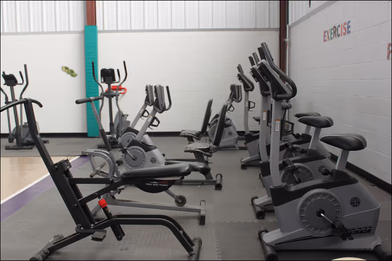 Indoor exercise room with multiple stationary bikes and exercise equipment arranged in rows on a padded floor. The walls are white with some colorful text and decorations, and the space is well-lit with natural light from windows above.