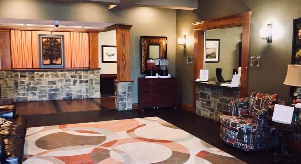 A cozy interior common area with a patterned rug on a wooden floor, a stone and wood reception desk with a chair behind it, a cushioned armchair with a colorful geometric pattern, a table lamp, framed artwork on the walls, and a wooden cabinet with beverage dispensers on top.