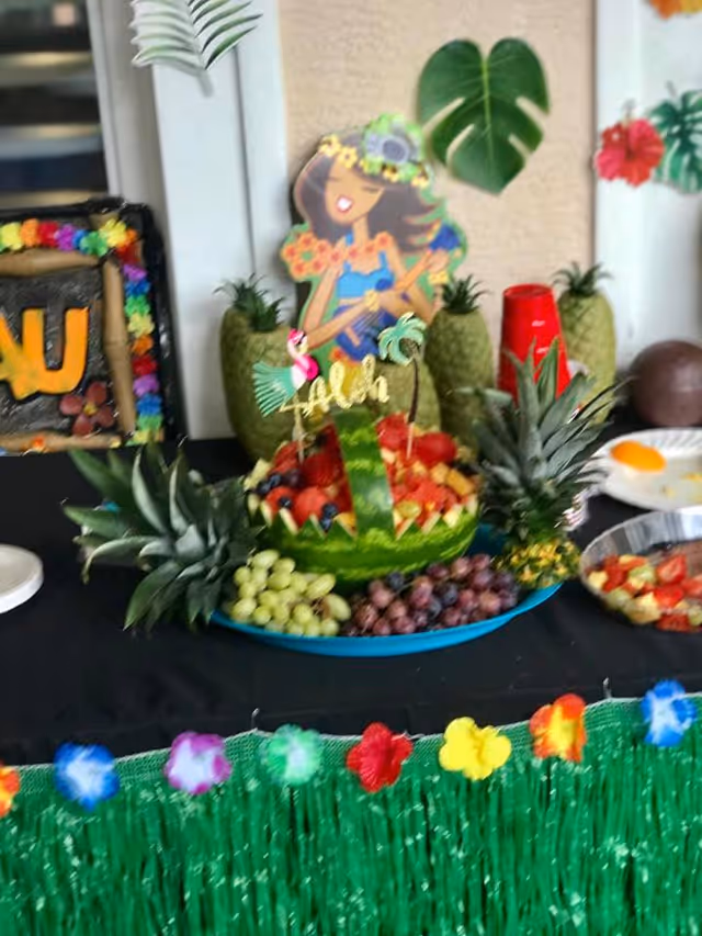 A tropical-themed fruit display on a table featuring a carved watermelon filled with various fruits, surrounded by pineapples and grapes. The table is decorated with a green grass skirt and colorful flower garlands. In the background, there is a cartoon cutout of a hula girl and some tropical leaves on the wall.