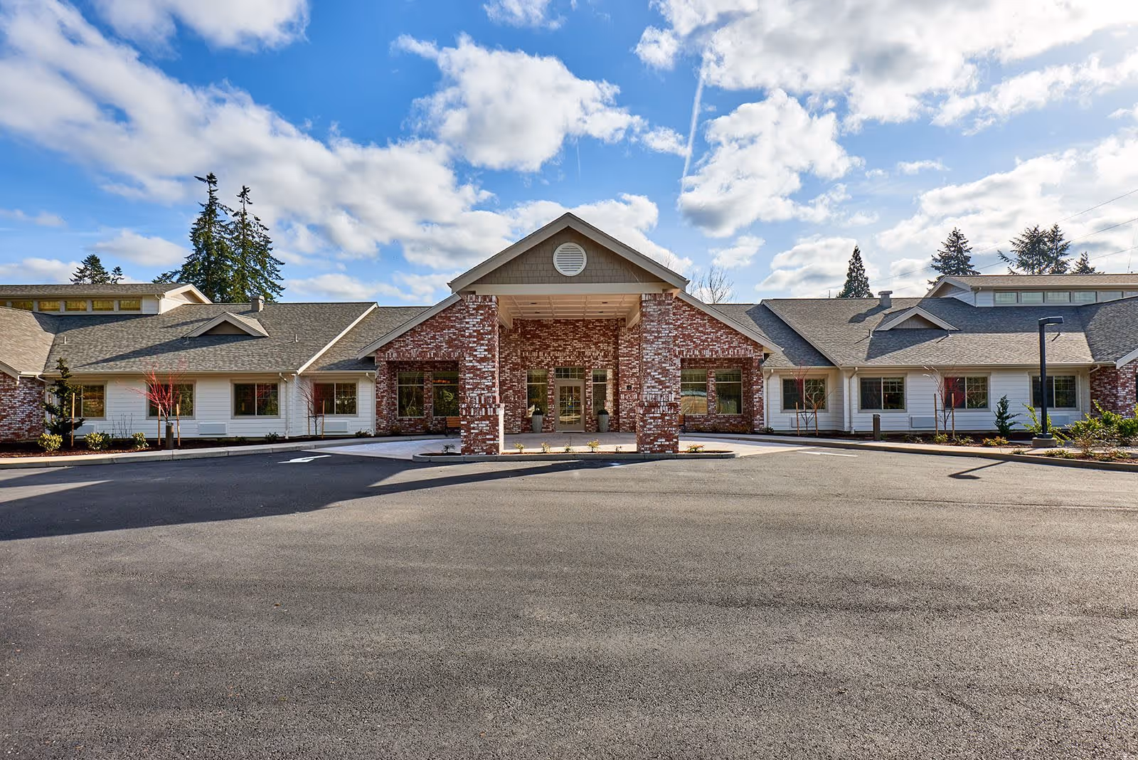 Front exterior view of The Rawlin at Riverbend Memory Care facility showing a large building with a covered entrance supported by brick columns, surrounded by a paved driveway and landscaped with small trees and shrubs under a partly cloudy sky.