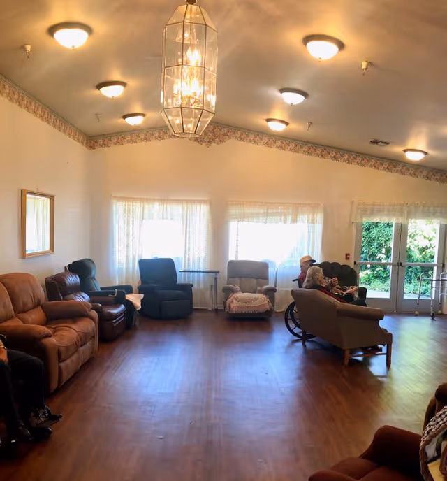 A spacious living room area in a senior care facility with multiple comfortable armchairs and sofas arranged around the room. Two elderly individuals are seated on a sofa near the windows, one in a wheelchair. The room has wooden flooring, white walls with a floral border near the ceiling, and several ceiling lights including a central chandelier. Large windows with sheer curtains allow natural light to enter, and a glass door leads outside to greenery.