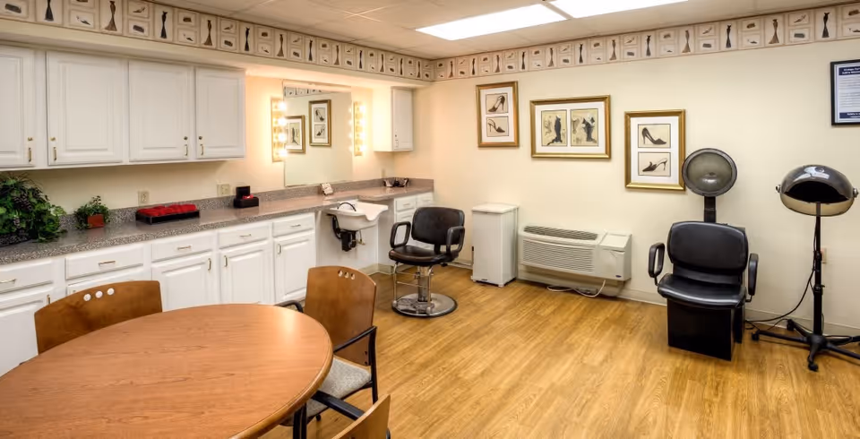 Interior of a salon room with wooden flooring, white cabinets along one wall, a round wooden table with chairs, two black salon chairs, a hair dryer, and framed pictures of shoes on the walls.