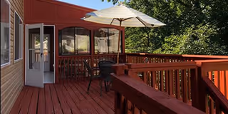 Outdoor wooden deck area with red railing and flooring, featuring a table with chairs and a large white patio umbrella, surrounded by green trees and adjacent to a building with beige siding and a red exterior section.