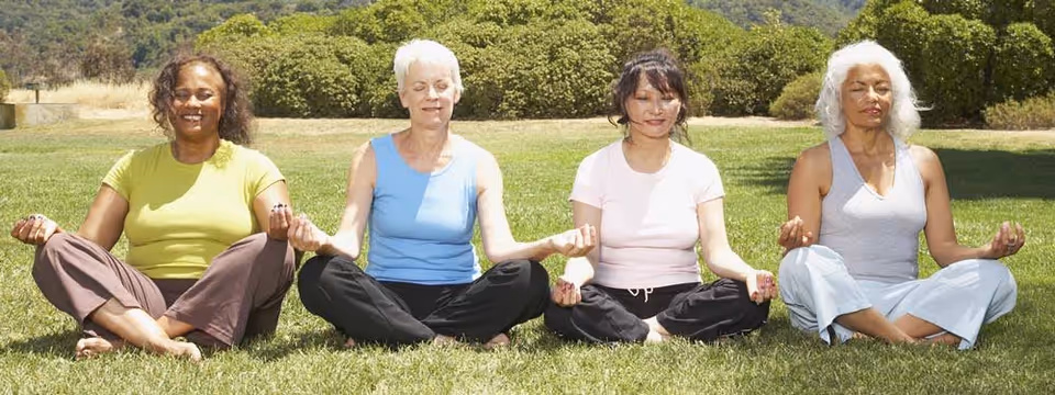 Four women sitting cross-legged on grass outdoors, meditating with eyes closed and hands resting on knees in a peaceful setting with bushes and hills in the background.