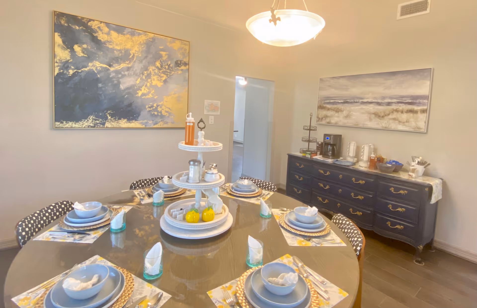 Round dining table set with place settings and a tiered centerpiece in a bright dining room with wall art and a buffet.