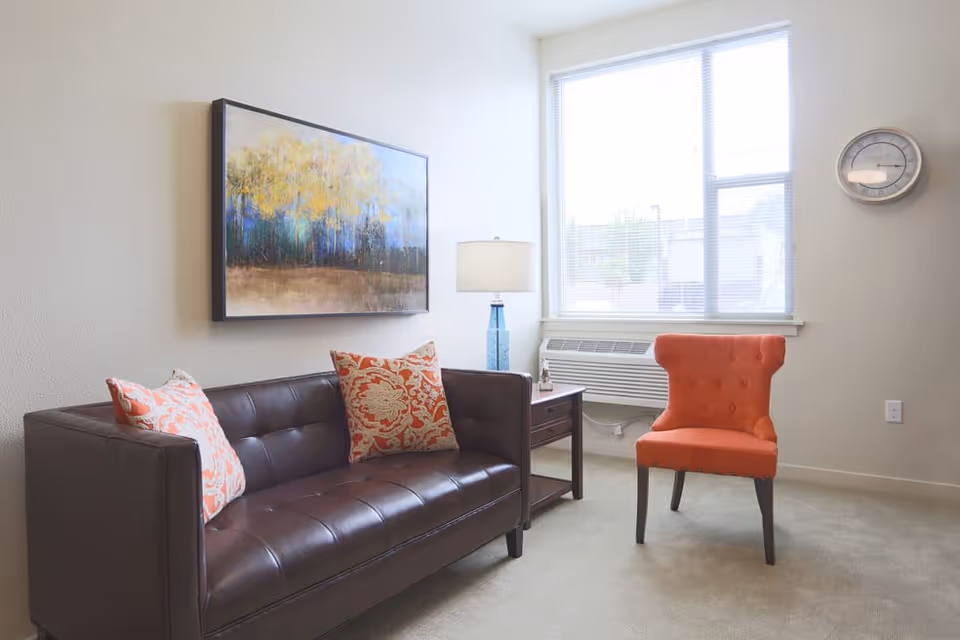 A bright living room with a dark brown leather sofa adorned with two patterned orange and white pillows, an orange upholstered chair, a wooden side table with a blue lamp, a large window with blinds, a wall clock, and a framed painting of trees on the wall.