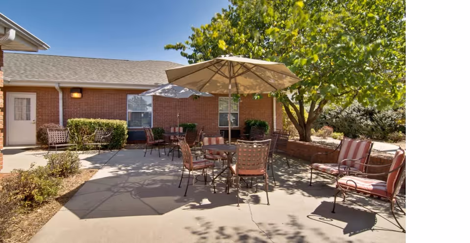 Outdoor patio area at Spring Ridge Assisted Living with several metal tables and chairs featuring red and white striped cushions. Large beige umbrellas provide shade, and there is a tree with green leaves casting shadows on the concrete ground. The background shows a brick building with windows and a door.