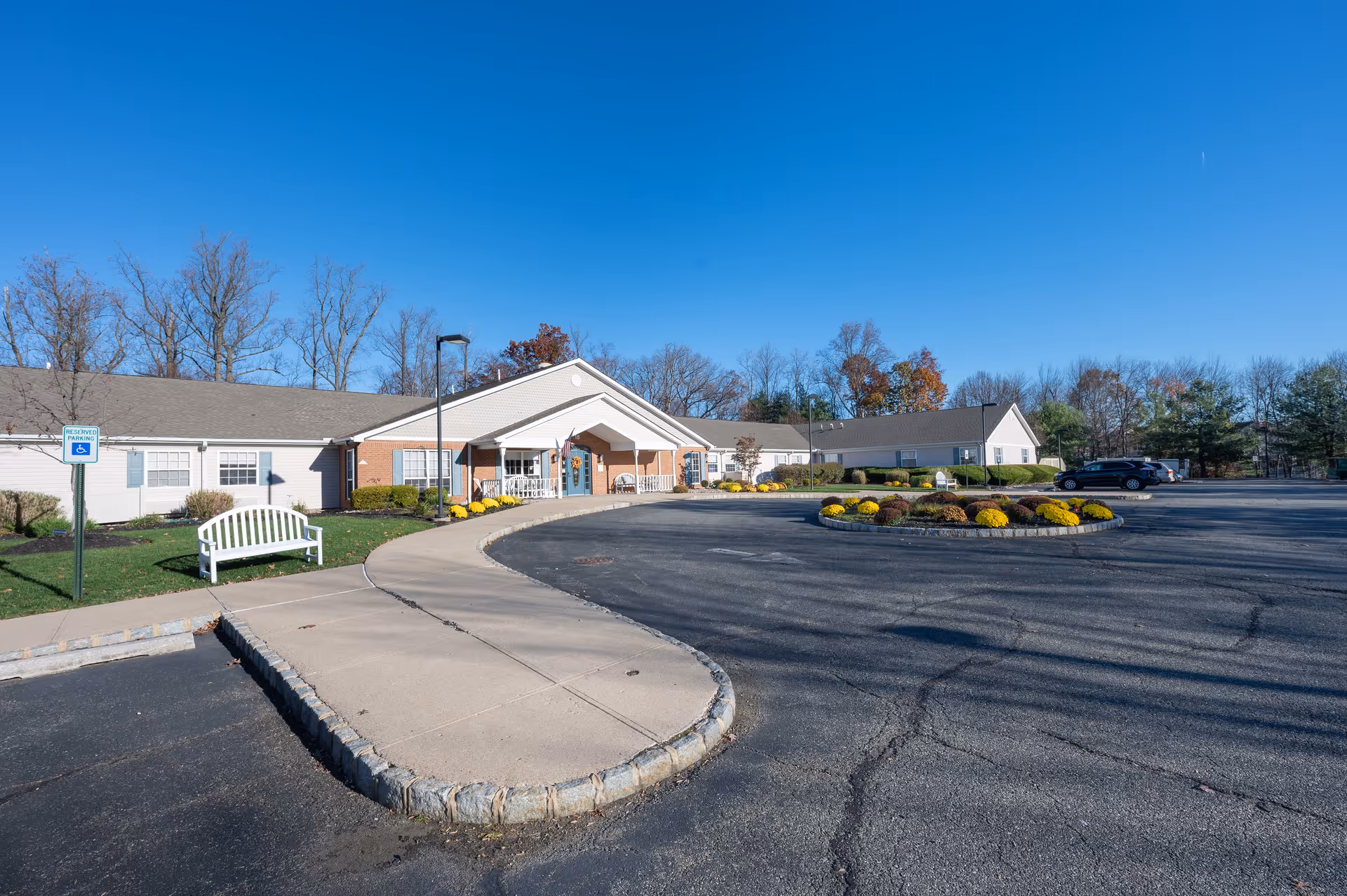 Exterior view of Arden Courts - ProMedica Memory Care Community in Whippany, showing a single-story building with a covered entrance, a circular driveway with a landscaped roundabout featuring yellow flowers, a white bench near a handicapped parking sign, and a clear blue sky.