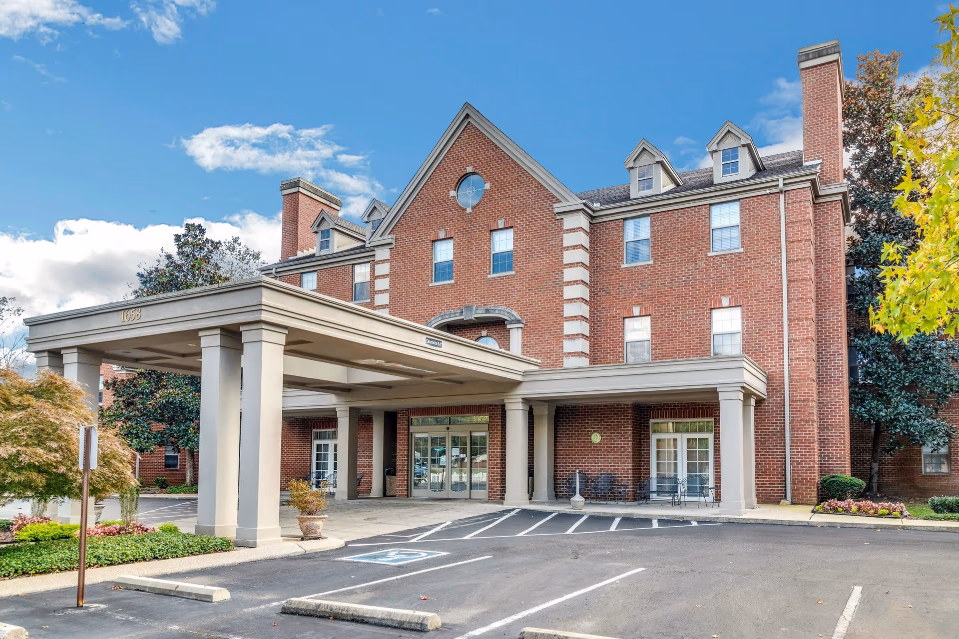 Front exterior view of a three-story brick building with a covered entrance supported by columns. There are several windows, a circular window near the roof peak, and a parking area with marked spaces including a handicapped spot. Trees and landscaping surround the building under a partly cloudy blue sky.