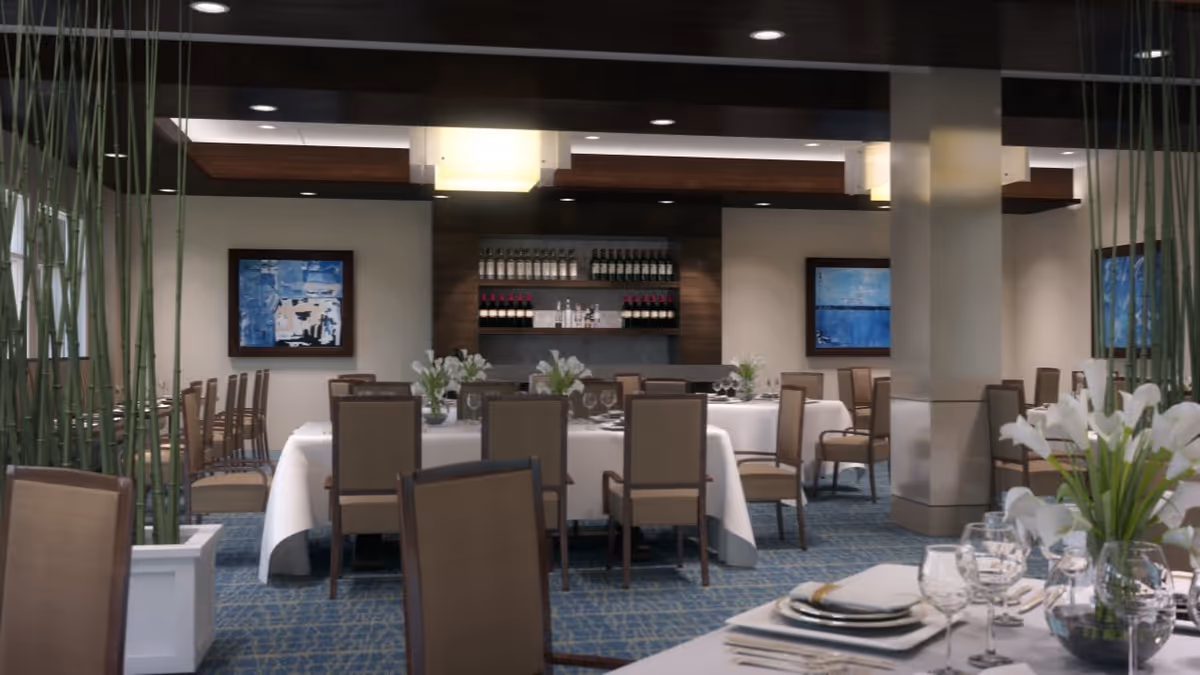 A well-lit dining room with multiple tables covered in white tablecloths, set with plates, glasses, and silverware. The room features beige cushioned chairs, blue patterned carpet, modern ceiling lights, and decorative plants. There are framed abstract paintings on the walls and shelves with bottles in the background.