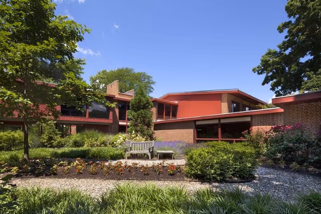 A landscaped garden area with a wooden bench and a small table in front of a brick building with red accents under a clear blue sky. The garden has various plants, flowers, and trees surrounding the seating area.