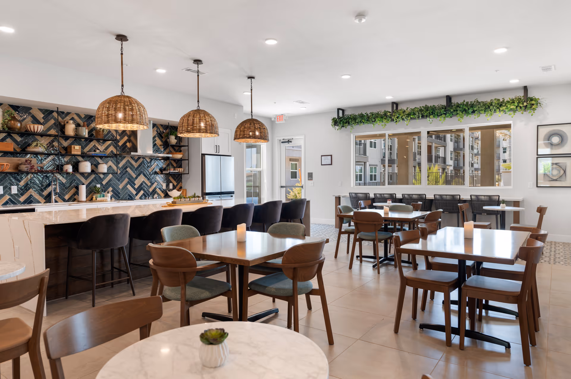 Bright communal dining area with multiple tables and chairs, a marble-topped bar with pendant lights, and decorative shelving and greenery.