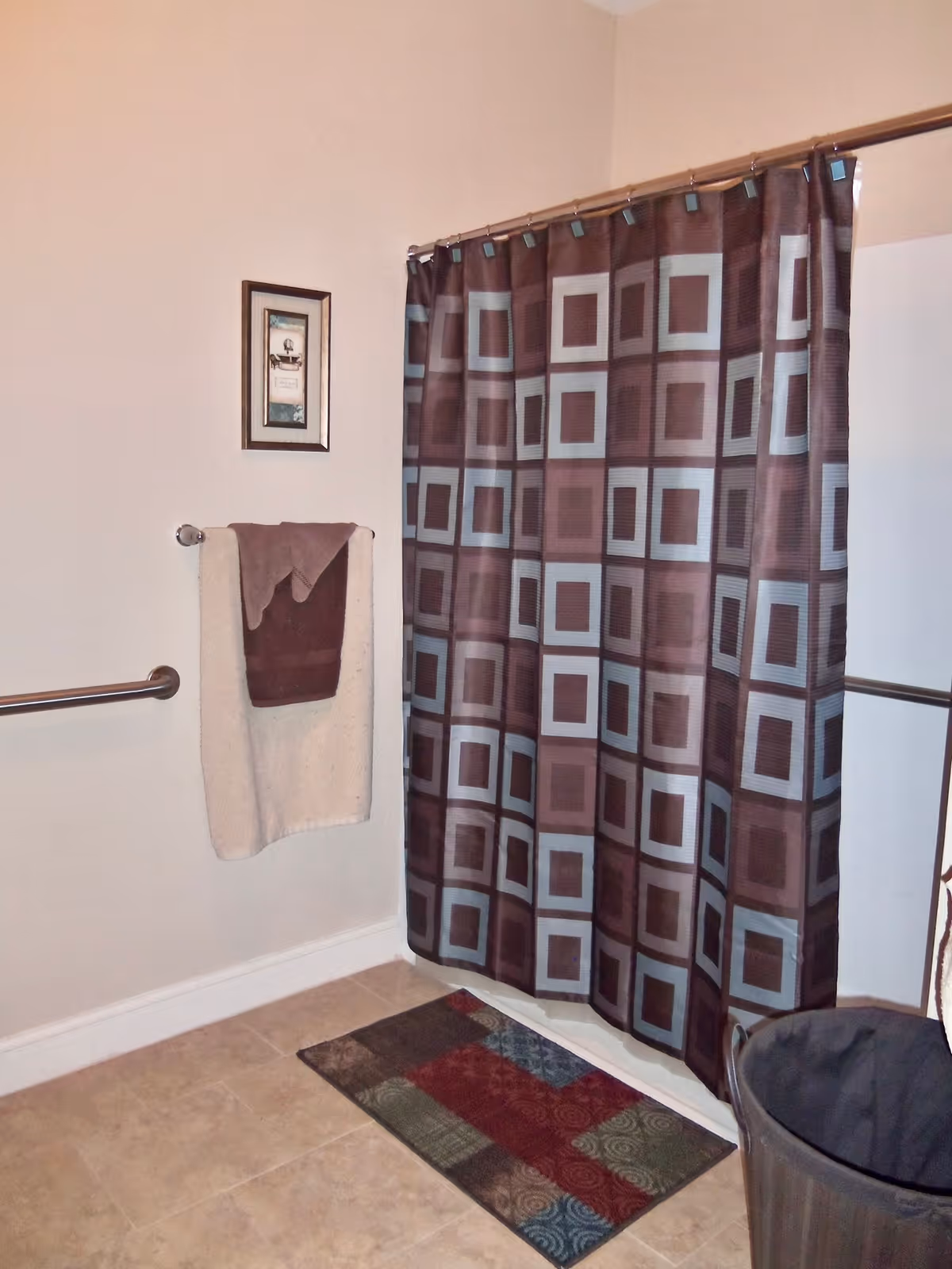 Bathroom interior with a patterned shower curtain, towel on a bar, grab rails, a bath mat, and a waste bin.