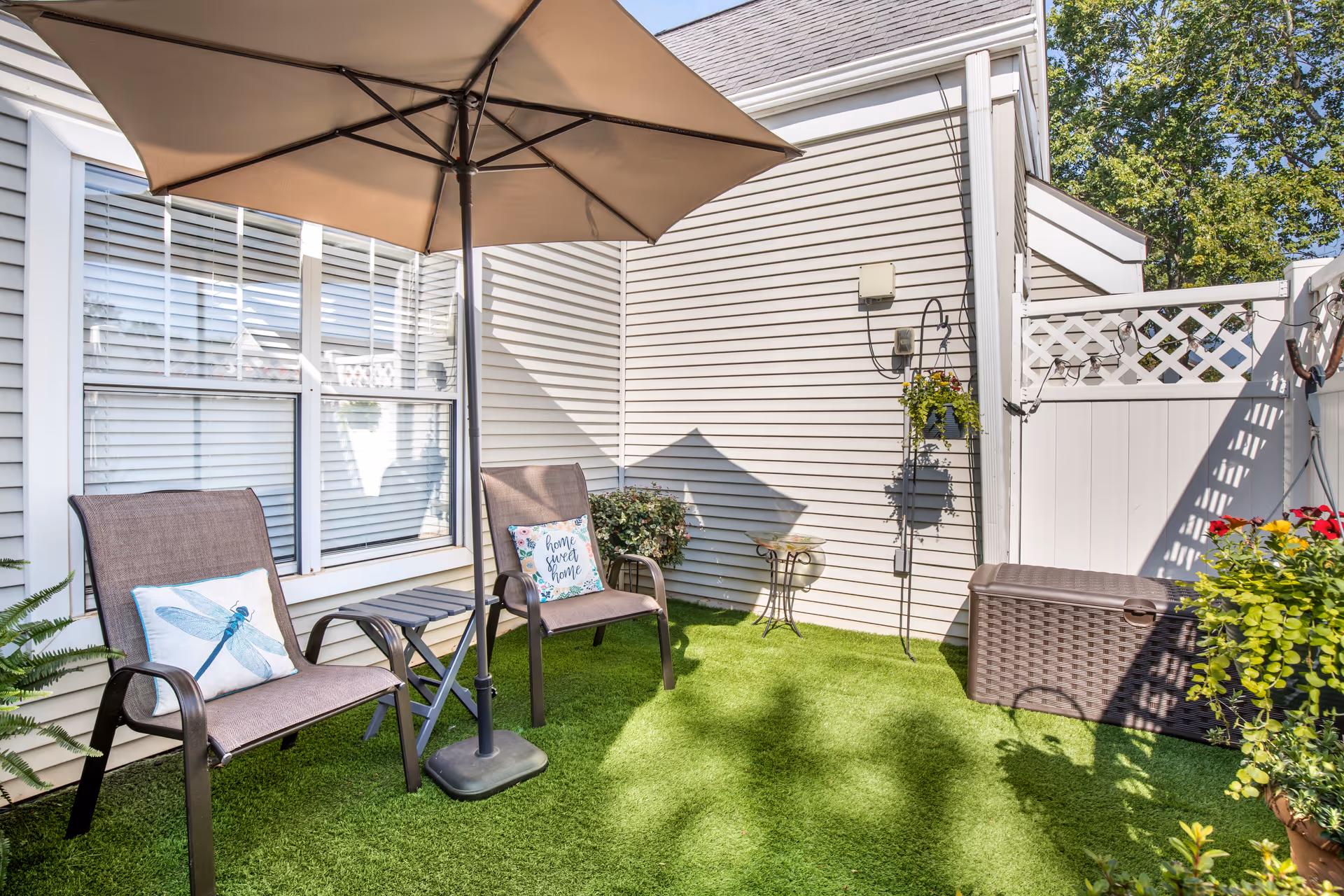 Small fenced patio with two chairs and cushions, a large umbrella, artificial turf, storage chest and potted plants beside a building.