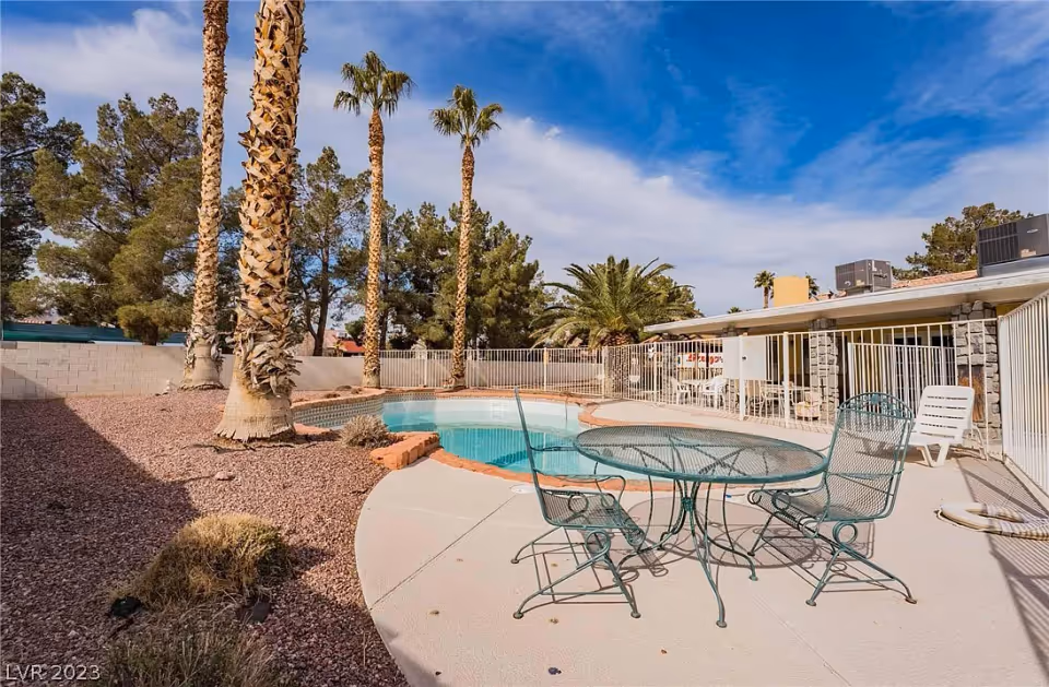 Outdoor pool and patio area with a metal table and chairs, lounge chairs, palm trees, and a fenced single-story building under a blue sky.