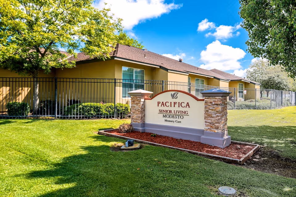 Exterior view of Pacifica Senior Living Modesto facility with a sign in the foreground displaying the facility name and 'Memory Care'. The building is beige with a brown roof, surrounded by green grass, trees, and a black metal fence under a blue sky with some clouds.