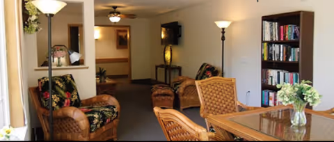 A cozy interior living area with wicker furniture including armchairs and a glass-top table with chairs. There is a bookshelf filled with books on the right side and a floor lamp next to it. The room has soft lighting with wall sconces and a ceiling fan light fixture. A hallway is visible in the background.