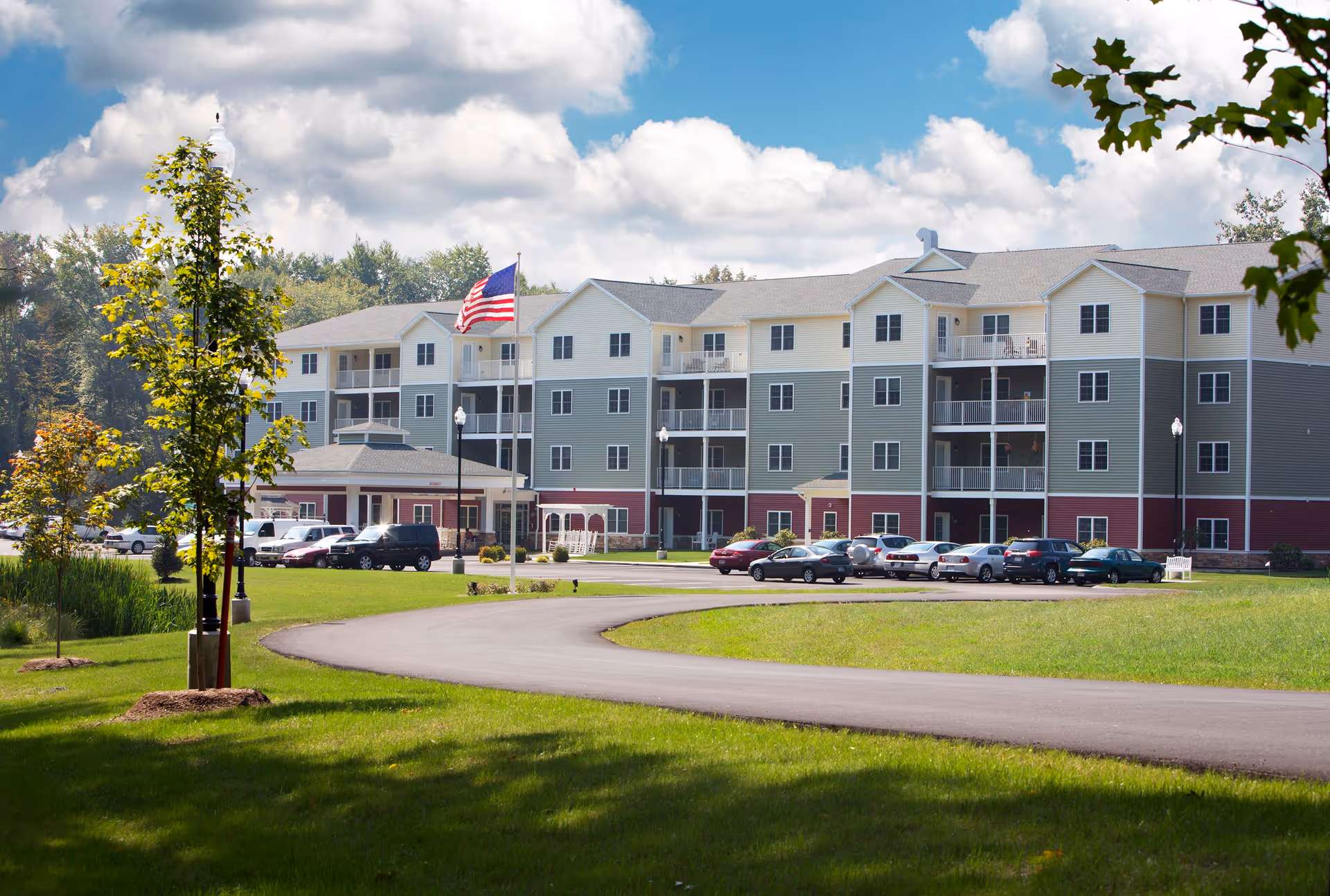 Four-story senior living building with balconies, an American flag on a flagpole, parked cars, and a curved driveway on landscaped grounds.