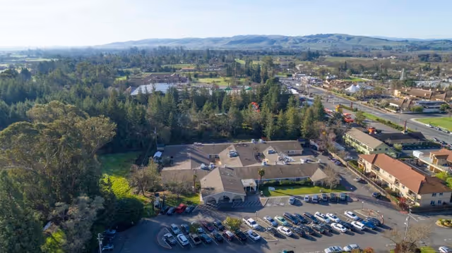 Aerial view of Broadway Villa Post Acute facility surrounded by trees and greenery, with a parking lot filled with cars in the foreground and a road with buildings in the background under a clear sky.