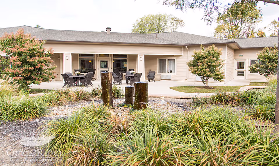 Outdoor patio area of Quail Ridge Assisted Living & Memory Care featuring several tables and chairs arranged for seating, surrounded by landscaped greenery and small trees, with a beige building in the background.