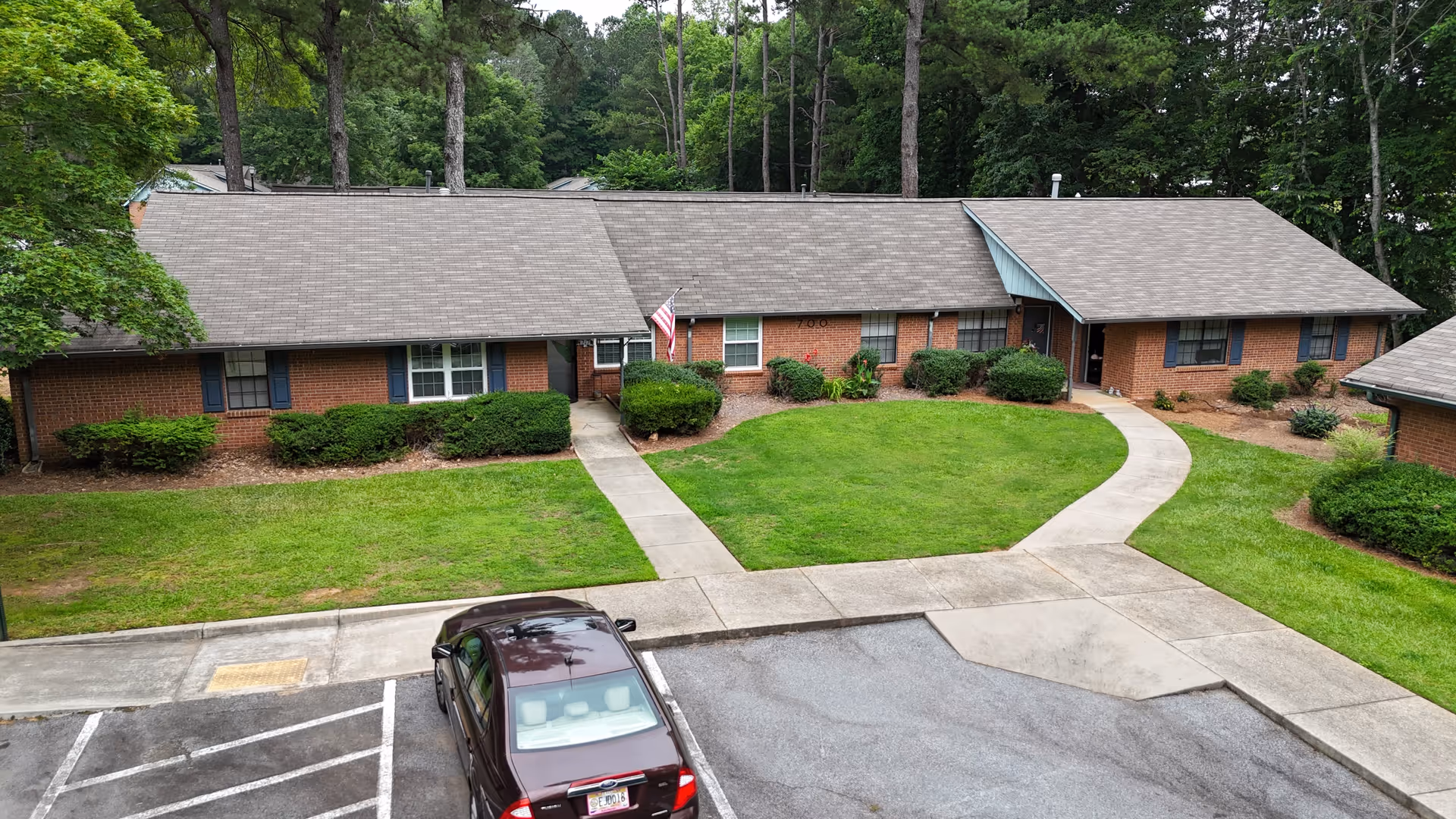 Exterior view of a single-story brick building with a gray shingled roof, surrounded by green grass, bushes, and tall trees in the background. A concrete walkway leads to the entrances, and a dark-colored car is parked in the parking lot in front of the building.