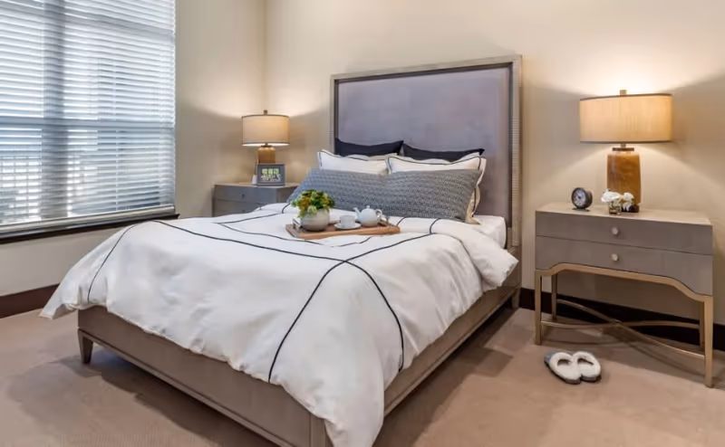 A neatly made bed with white bedding featuring black lines in a bedroom. The bed has a gray upholstered headboard and multiple pillows. There are two matching nightstands on either side of the bed, each with a lamp. One nightstand has a small clock and a white flower decoration, while the other has a framed photo and a small plant. A pair of white slippers is placed on the carpeted floor near one nightstand. A window with closed blinds is visible on the left side of the room.